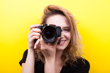 Beautiful woman holds a camera at her eye taking a picture on yellow background in studio photoの写真素材