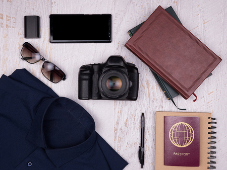Flat lay top view of traveler accessories on white wooden background. Digital camera, shirt, books, smartphone, paper notebook with pen, sunglasses and passportの写真素材