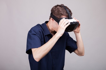 Young man in casual blue shirt wearing a VR virtual reality headset on gray background in studio photoの写真素材