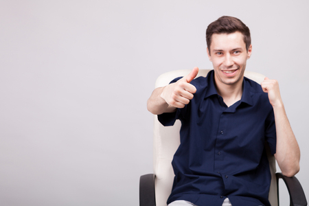 Young businessman in casual blue shirt showing thumbs up to the camera while sitting in an office chair over gray background in studio photoの写真素材