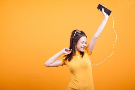 Cute girl dancing while listening music on her phone in studio on yellow backgroundの写真素材
