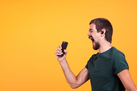Cool joyful young man listening and enjoying music in his headphones while holding his smartphone in...