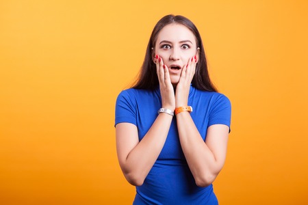Surprised and amazed girl holds her cheecks in studio over yellow background