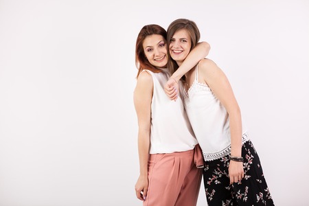 Two beautiful girl friends embracing each other on white wall in studio photoの写真素材