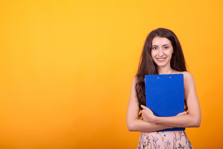 Attractive young girl holding blue clipboard in studio over yellow background. Successful young...