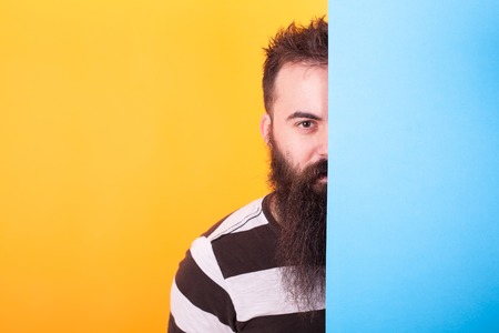 Handsome bearded hipster hiding behind a blue panel over yellow backgroundの写真素材