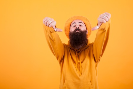 Portrait of bearded hipster with hat showing thumb down sign over yellow backgroundの写真素材