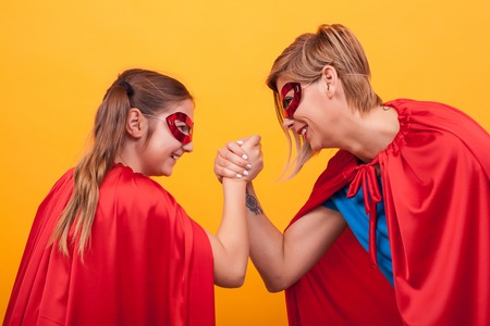 Mother and daughter dressed like superheroes playing arm wrestling over yellow background.の写真素材