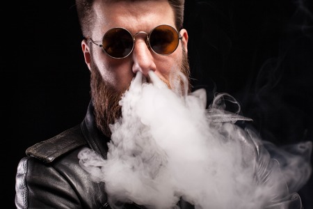 Attractive bearded man exhaling cigarette smoke on his nose over black background. Man portrait. Stylish beard.の写真素材
