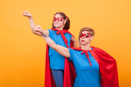 Mother and daughter dressed like heroins holding their fist in the air and looking away over yellow background. Happy childhood. Mom super powers.の写真素材