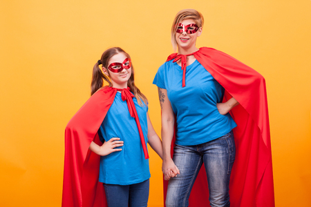 Little girl and mother dressed like superheros holding hands and smiling to the camera over yellow background. Super powers. Happy kid.の写真素材