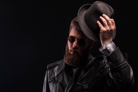 Misterious handsome bearded man with half of his face in the shadow while holding his hat over black background. Attractive man.の写真素材