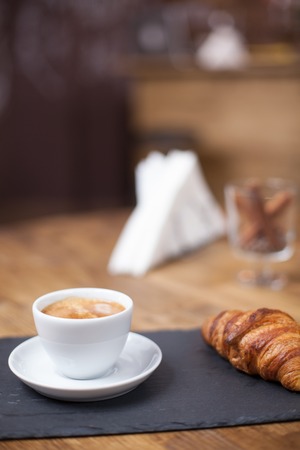 Close up of cup of coffee served with croissant in a cozy coffee shop. Coffee aroma.の写真素材
