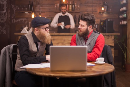 Two handsome businessmen looking at each other while talking about finances in a vintage pub. Stylish beard. Coffee cup. Laptopの写真素材