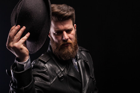 Low key portrait of handsome man holding stylish hat over black background. Bearded man. Dramatic portrait.の写真素材