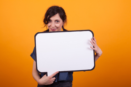 Portrait of beautiful young woman holding a white thoughts bubble in studio over yellow background. Speech space available. Happy woman with blank sign.の写真素材