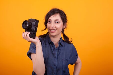 Portrait of young beautiful woman smiling at the camera and holding photo camera in studio over yellow background. Gorgeous young photographer woman with camera.の写真素材