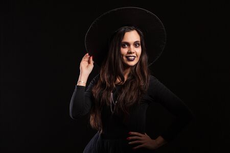 Beautiful young brunette witch holding her hat with her hand over black background. Make up for...
