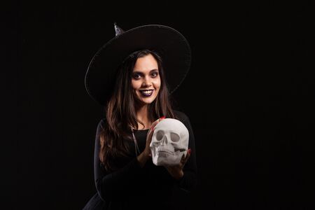 Malefic witch woman with a big hat looking into the camera with a human skull in her hands. Pretty young girl dressed up like a witch for halloweenの写真素材