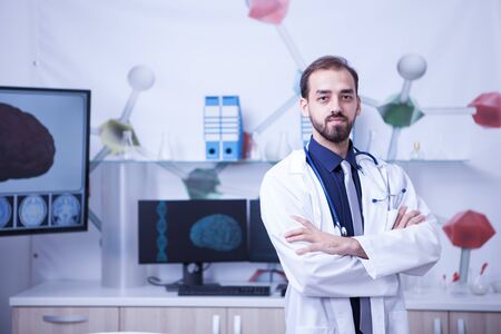 Attractive bearded doctor with arms crossed looking at the camera in his hospital cabinet. Young surgeon in the cabinet.の写真素材