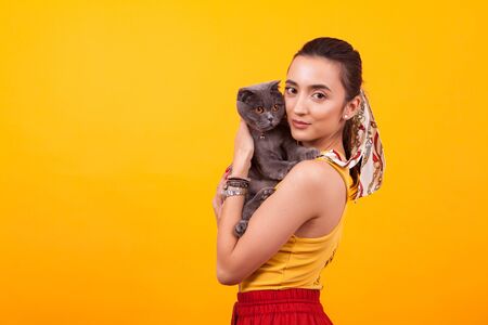 Beautiful young girl holding her adorable cat and looking at the camera in studio over yellow background. Girl and cat friendship.の写真素材