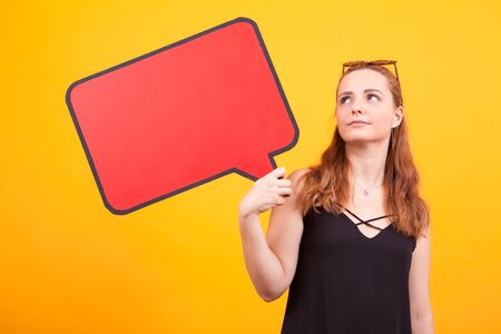 Thoughtful young woman holding an empty bubble in studio over yellow background.