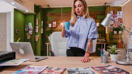 Young designer drinking coffee and looking at a table on which some prints are arranged. Loft office