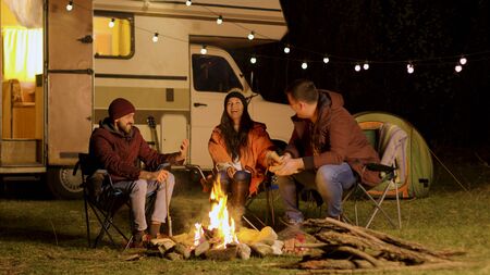 Group of close friends laughing together around camp fire. Retro camper van. Light bulbs in the background.の写真素材
