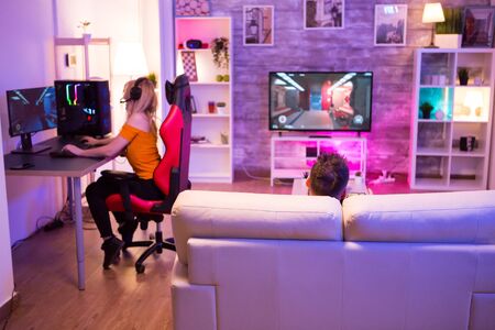 Back view of man sitting on sofa and playing games. Female sitting on gaming chair playing on computer.の写真素材
