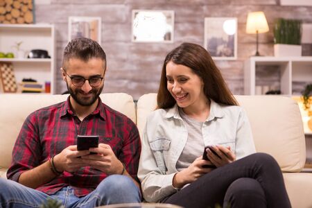 Cheerful young couple laughing together while sitting on sofa. Boyfriend showing something to his...