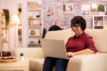 Old elderly woman on her sofa working on a modern laptop in her cozy living room. Her husband is in the backgroundの写真素材