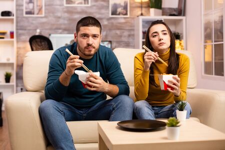 In modern cozy living room couple is enjoying takeaway noodles while watching TV comfortably on the sofaの写真素材