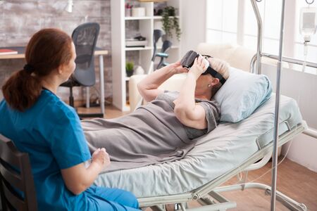 Caucasian old woman laying in bed in nursing home using virtual reality glasses. Female doctor.の写真素材