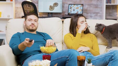 Bearded man eating chips and using remote control to change tv channels while his girlfriend is playing with the cat.の写真素材