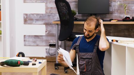 Confused worker rubbing his head while reading instruction for new furniture. Electric Drill on...