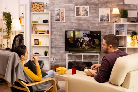 Back view of couple in living room watching a movie on the TV while eating takeaway foodの写真素材