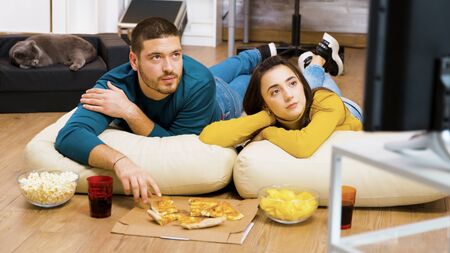Attractive couple relaxing on pillows for the floor watching tv and eating pizza with their cat sleeping in the background.の写真素材