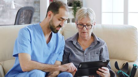 Male assistant teaching senior retired elderly old woman how to use her tablet computer in nursing...
