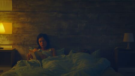 Beautiful wife reading a book in a dark room while his husband is sleeping. Lamp turned on.の写真素材