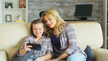 Young mother and her daughter relaxing on the couch watching a funny video on smartphone.の写真素材