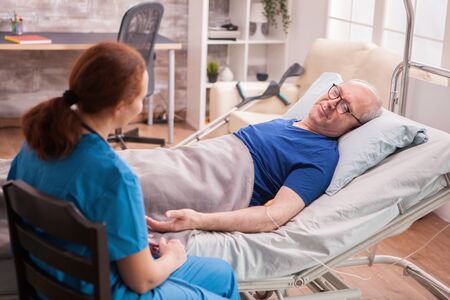 Back view female doctor in nursing home sitting next to old man lying in bed.の写真素材