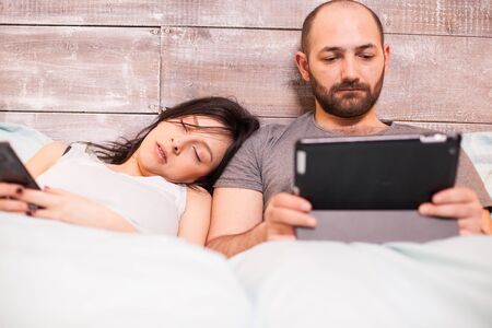 Beautiful wife in pajamas sleeping while her husband is working on tablet computer.の写真素材