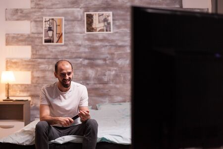 Cheerful man holding remote control watching tv at night.の写真素材