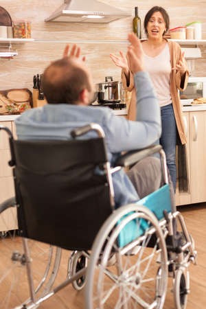 Wife screaming in kitchen to her disabled man in wheelchair while having a disagreement. Disabled paralyzed handicapped man with walking disability integrating after an accident.の写真素材