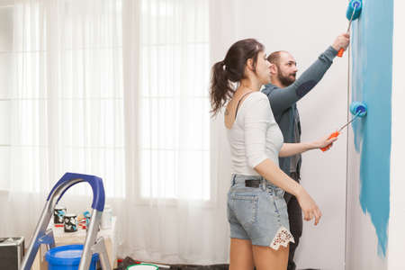 Caucasian married couple doing a makeover of their apartment painting the walls with roller brush....