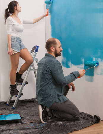 Tired young couple sitting on the floor covered with plastic wrap and painting the wall. Apartment redecoration and home construction while renovating and improving. Repair and decoratingの写真素材