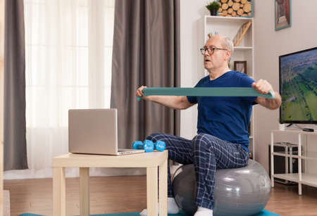 Strong old man training online with resistance band. Old person pensioner online internet exercise training at home sport activity with dumbbell, resistance band, swiss ball at elderly retirement age.の写真素材