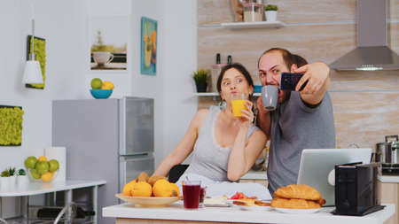 Cheerful young couple taking selfies during delicious breakfast in kitchen. Joyful married husband and wife making funny faces while taking a photo during breakfast in kitchen.の写真素材