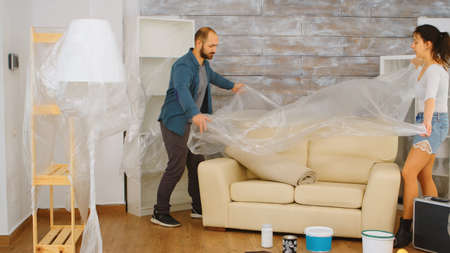 Couple wrapping sofa in plastic foil for protection while they are renovate living room. Home renovation, construction, painting work.の写真素材