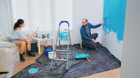 Husband changing wall paint using roller brush. Wife using paint brush. Apartment makeover. Couple...